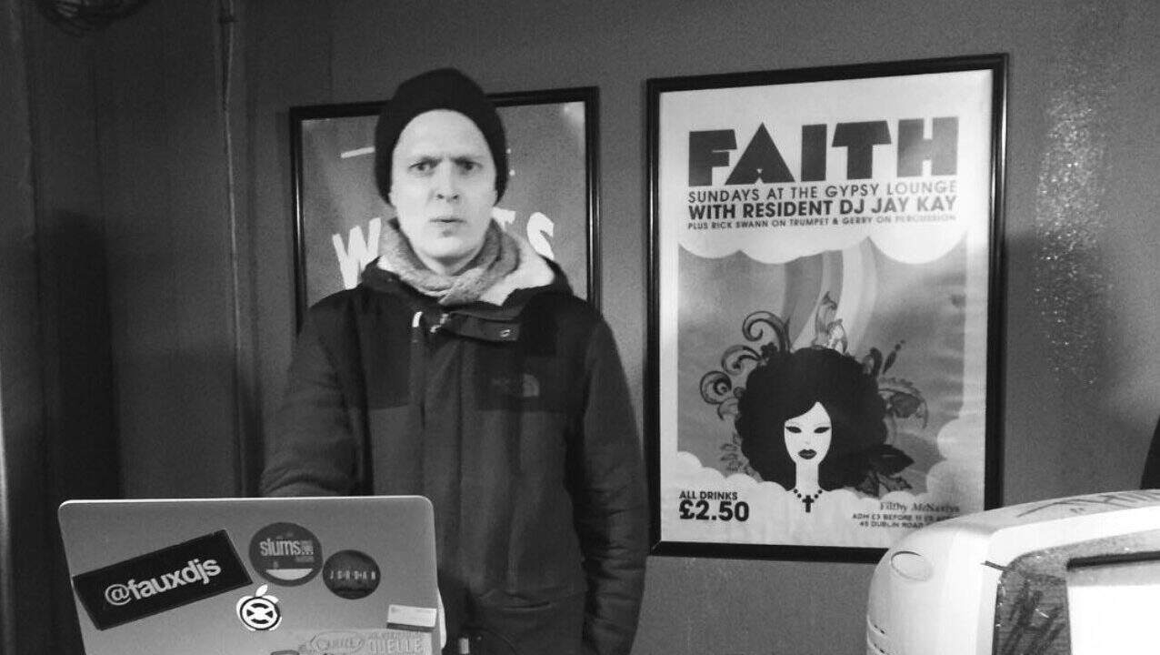 Young man in a black beanie and jacket standing in front of a laptop at Lavery's pub, Ireland, with Gypsy Lounge poster in the background, promoting a faith-themed event.