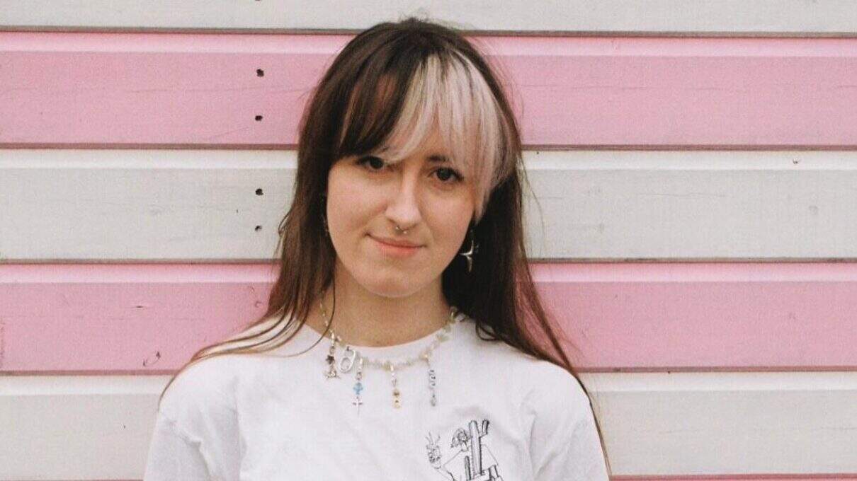 Sustainable fashion young woman with multi-coloured hair and piercings standing against pink and white wooden wall at Lavery's trendy boutique.