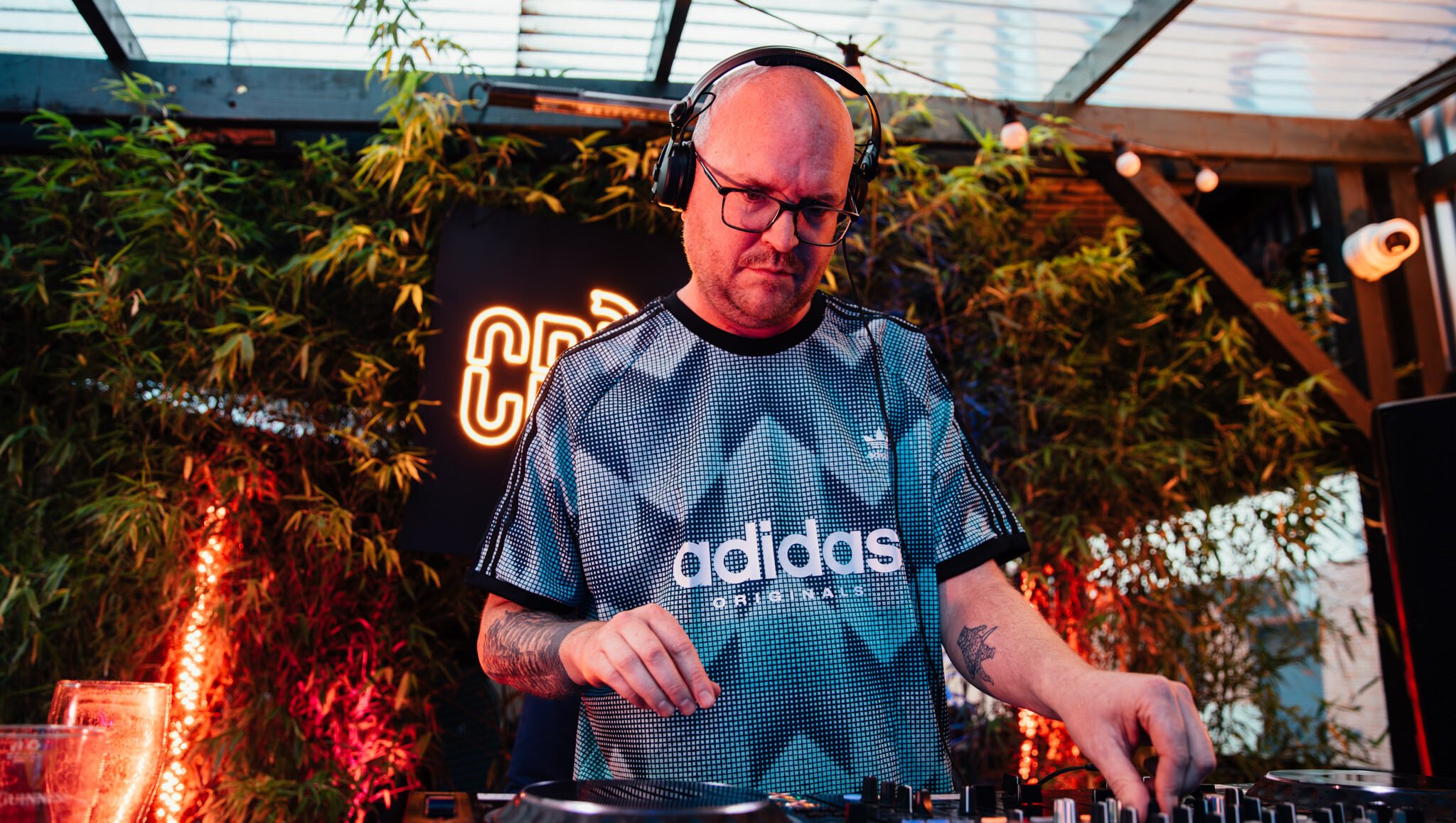 A male DJ wearing headphones and a sporty adidas shirt mixes music with professional DJ equipment in a vibrant club setting.