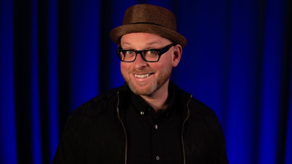 A smiling male comedian wearing glasses and a brown hat on stage at Lavery's Comedy Club, with a dark blue curtain background, promoting upcoming comedy events.
