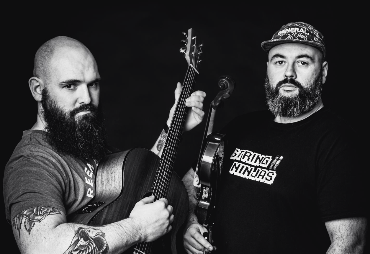 Two male musicians from the String Ninjas band performing live at Lavery's, with one playing an acoustic guitar and the other holding a violin, captured in black and white.