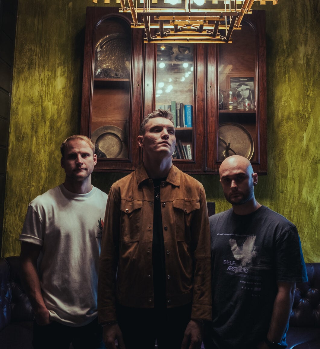 Three band members of Lavery's standing in a vintage room with green walls, wooden cabinet, and warm lighting, showcasing their musical style and presence.