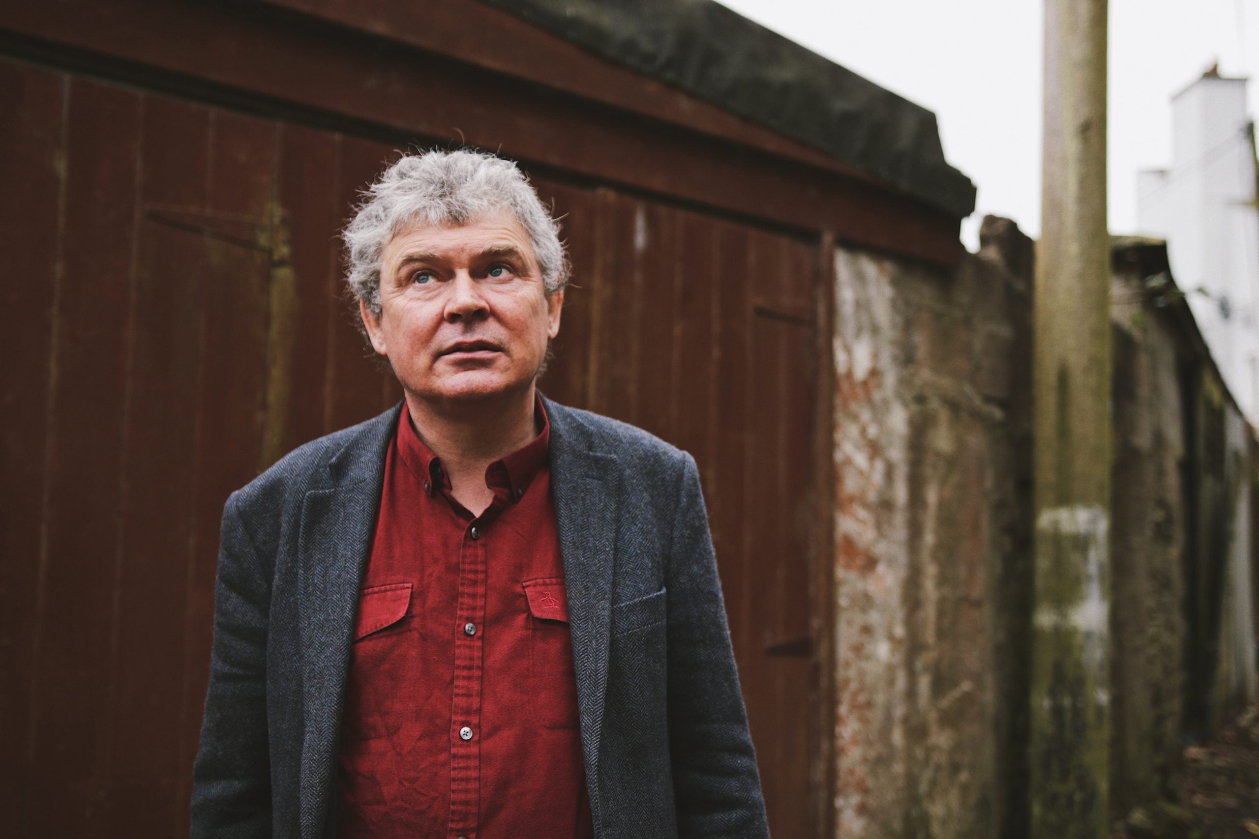 Portrait of a man with grey curly hair wearing a red shirt and dark blazer, standing outdoors near a rustic wooden shed, with a serious expression.
